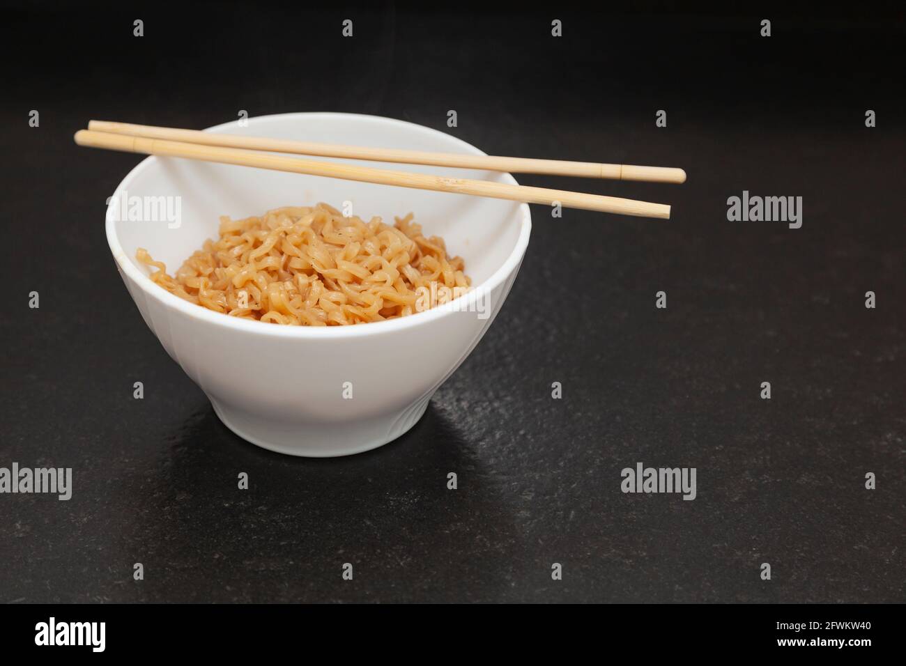 A kitchen bowl with ready-to-eat Chinese noodles. On the bowl are some oriental chopsticks. It is placed on a black granite bench in a kitchen and is Stock Photo