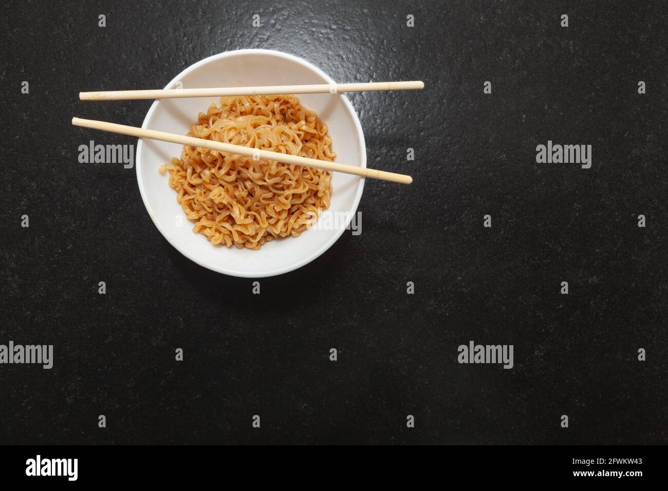 A kitchen bowl with ready-to-eat Chinese noodles. On the bowl are some oriental chopsticks. It is set on a black granite bench in a kitchen and viewed Stock Photo