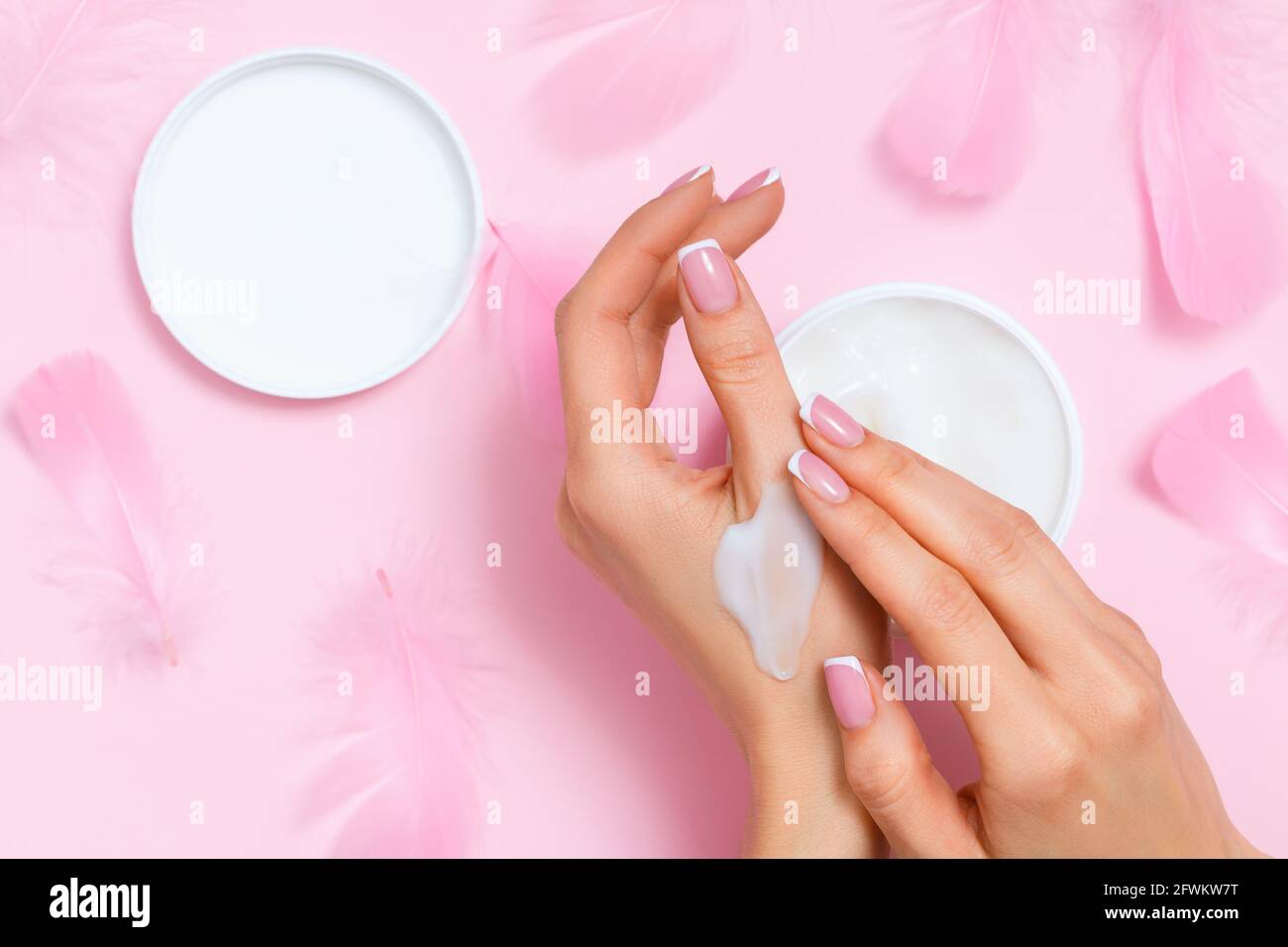 Top view of well-groomed female hands with moisturizing skin care cream in an open jar on a pink background with feathers. Copy space, close up, flat Stock Photo