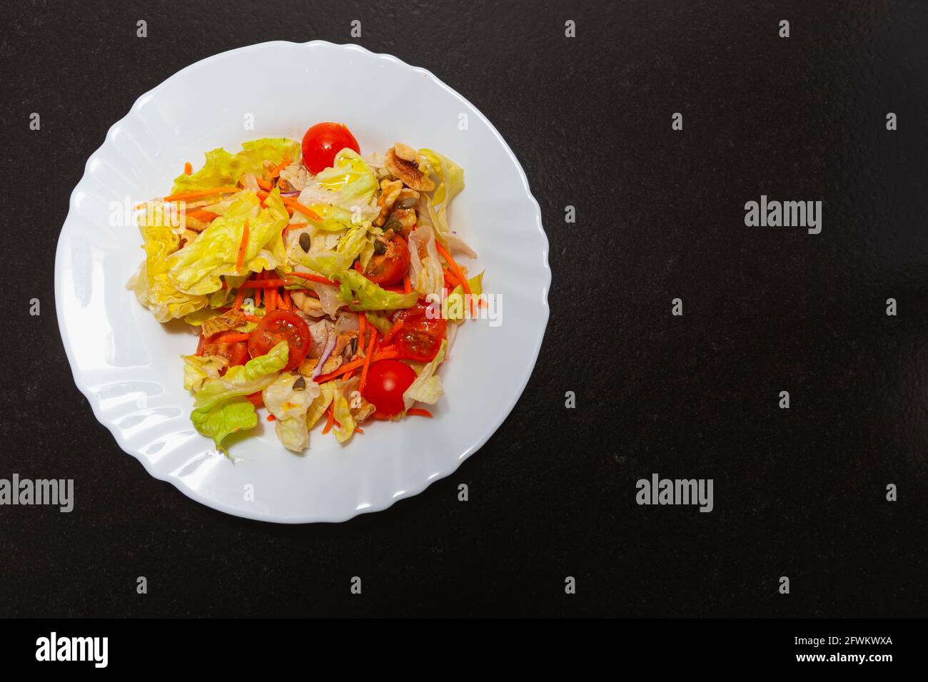 Combination of vegetables and greens in a ceramic plate on a dark bench in a kitchen. The recipe calls for lettuce, cherry tomato, carrot, walnuts, on Stock Photo