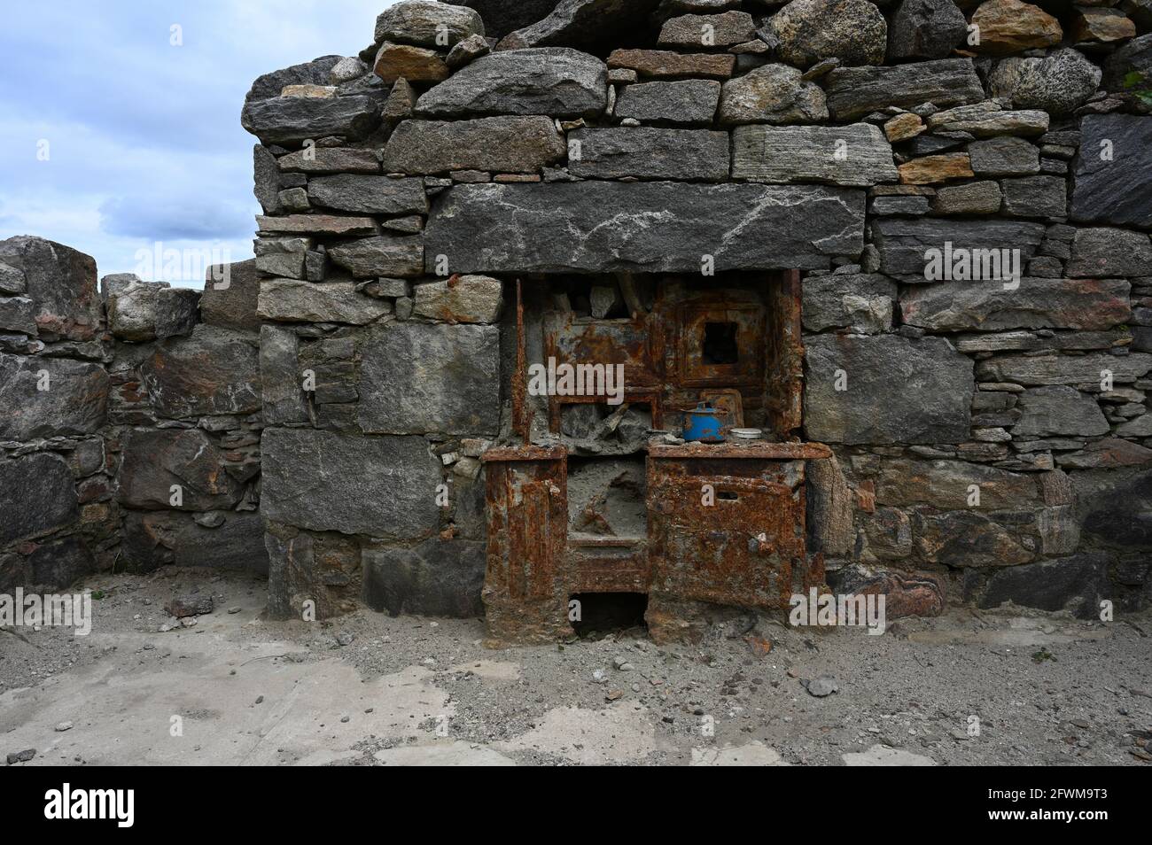 Old rusted cooking range and blue pot in ruins of stone cottage. Wall and roof are missing, showing blue sky. Exposed to elements. Isle of Lewis. Stock Photo