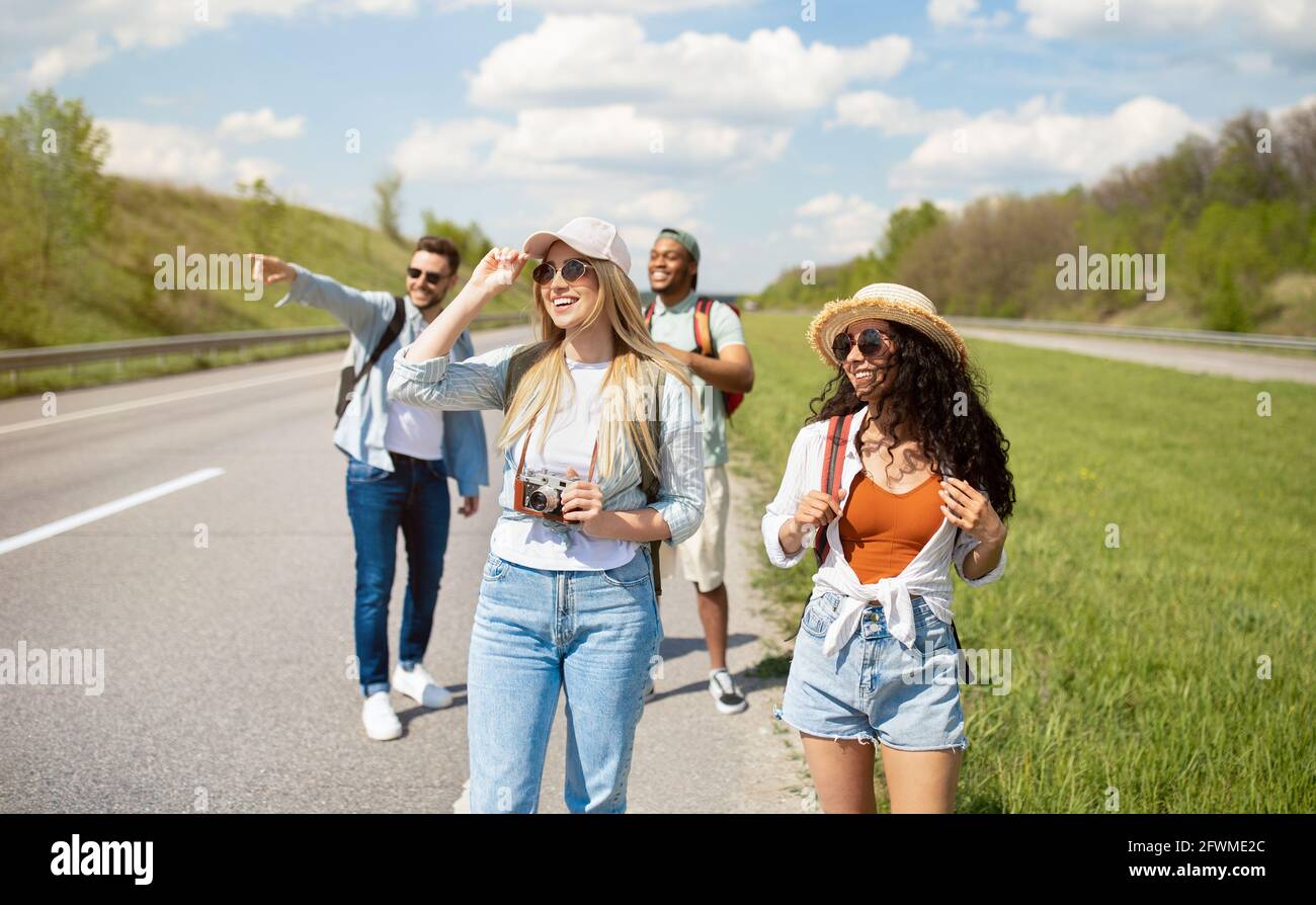 Group of millennial multiracial friends with backpacks walking along road, traveling together outdoors Stock Photo