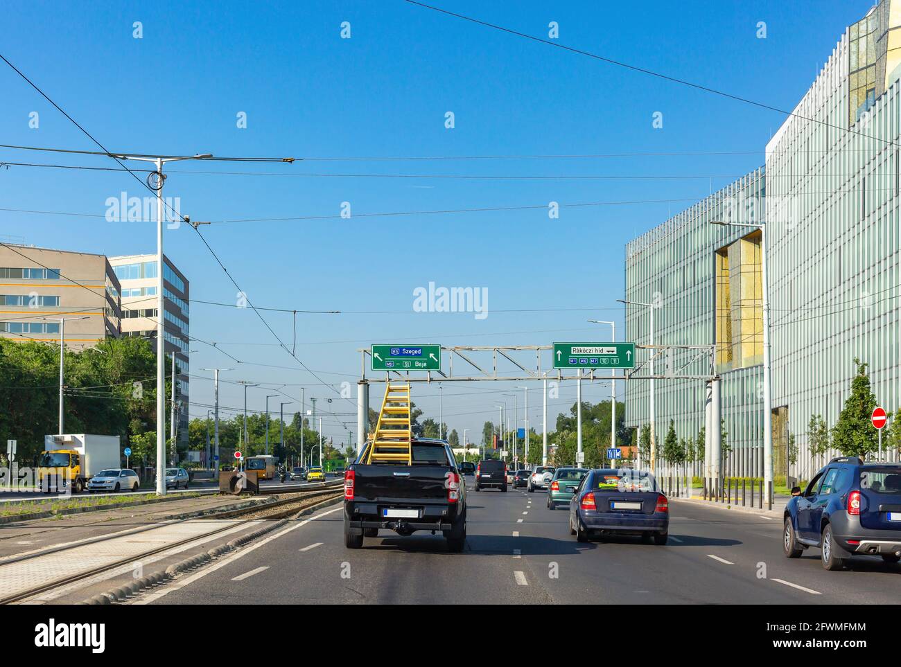 View of city. Cars on street, tram rails. Yellow ladder in cargo area of passenger truck. Back view. On sides of street are highrise buildings Stock Photo