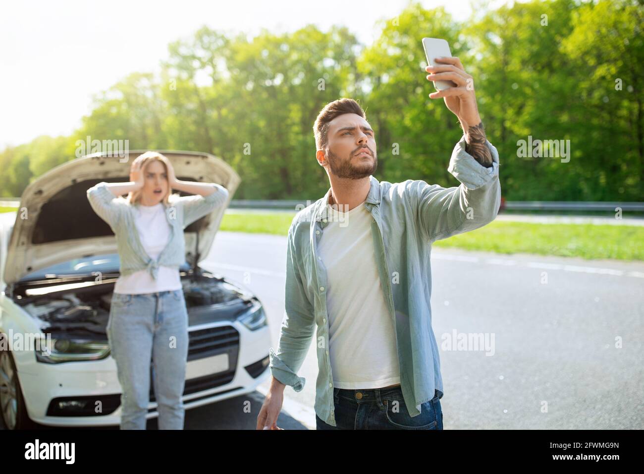 Sad young man standing on road next to broken car together with girlfriend, trying to call assistance. No connection Stock Photo