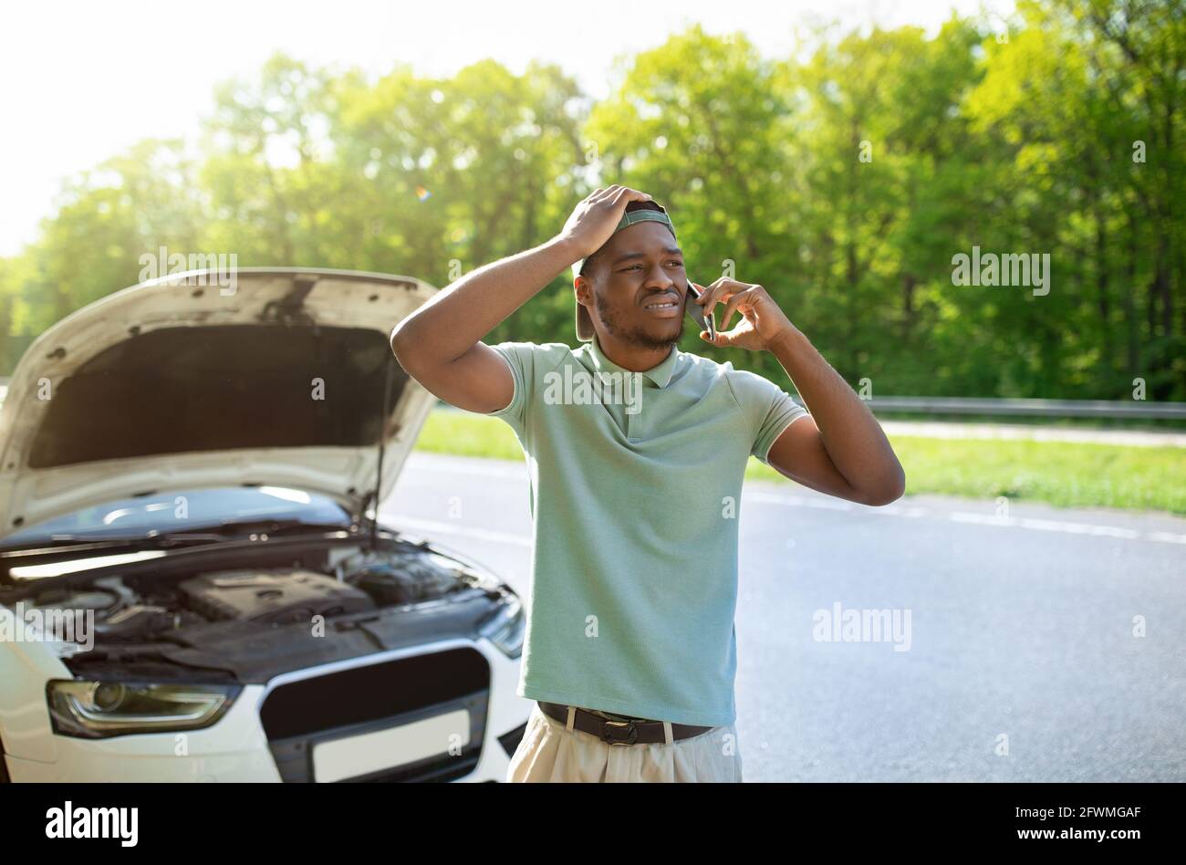 Unhappy black man standing near his broken car with open hood, calling breakdown road service on smartphone Stock Photo