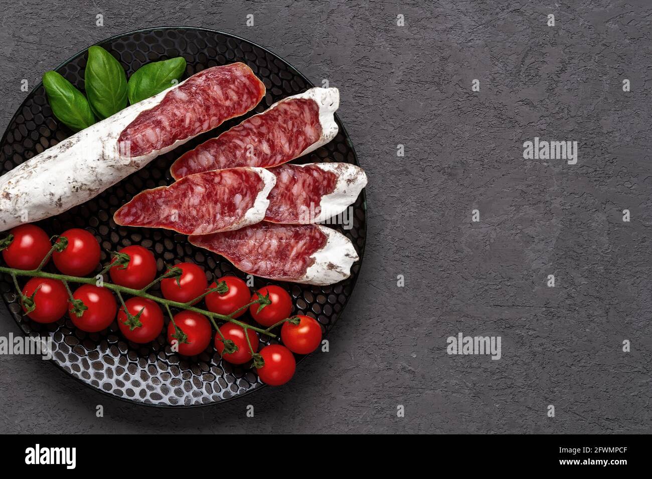 Sliced thin catalan fuet sausage and bunch of red cherry tomatoes on a black plate over dark rough surface. Traditional spanish dry-cured pork salami. Stock Photo