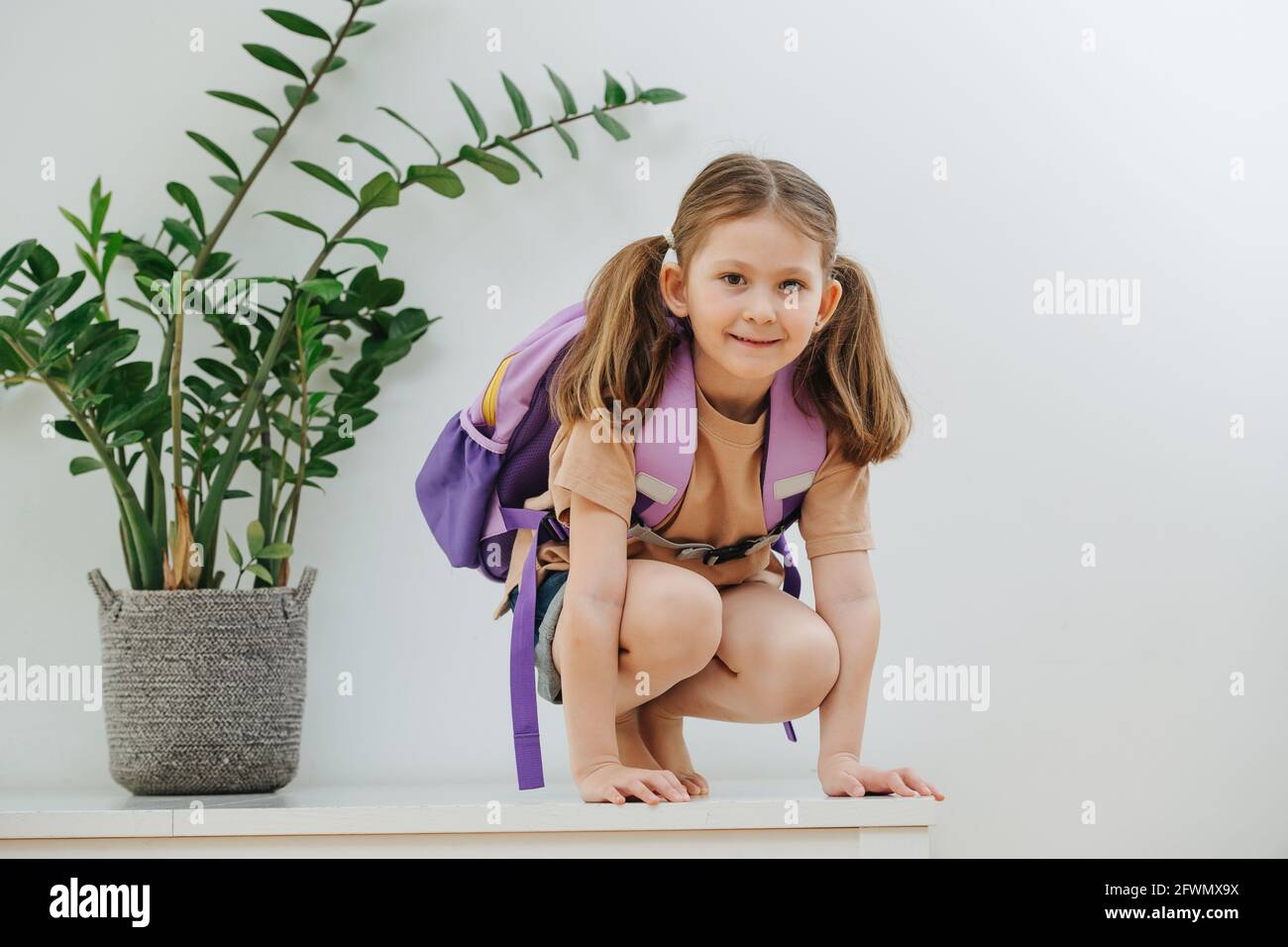 Playful little schoolgirl on a white table with purple backpack. Over white Stock Photo