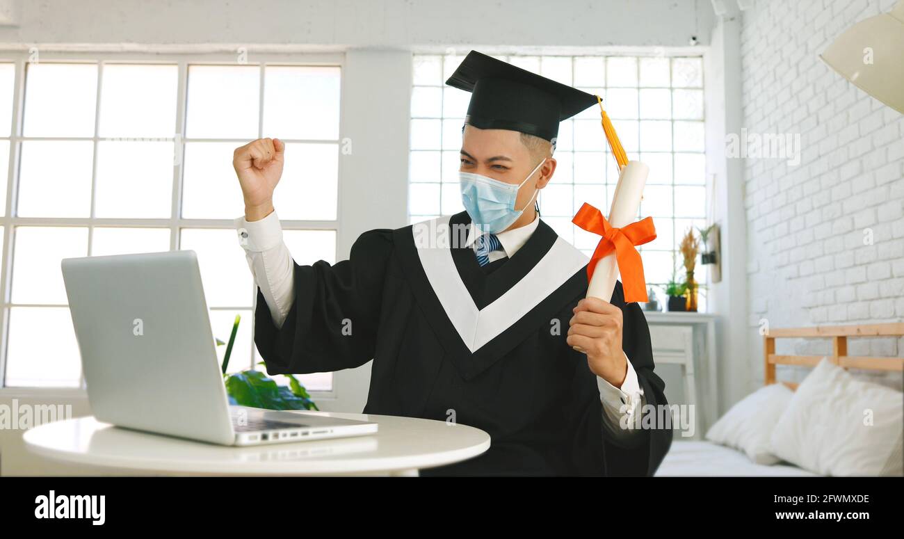 Happy excited college or university student wearing medical mask and sitting at desk with laptop, holding diploma and celebrating success Stock Photo