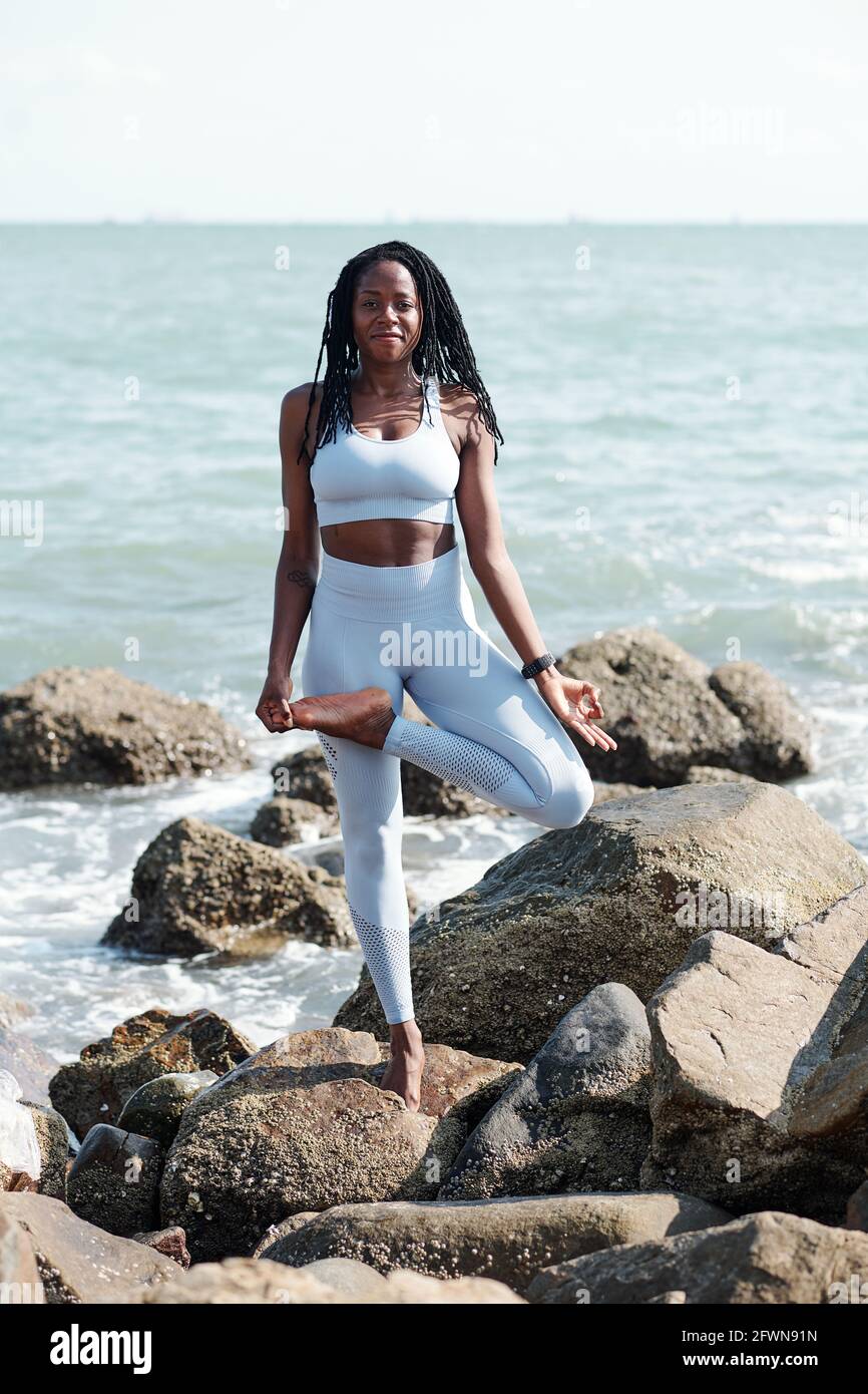 Smiling pretty fit young woman standing in tree pose on rocky beach ...