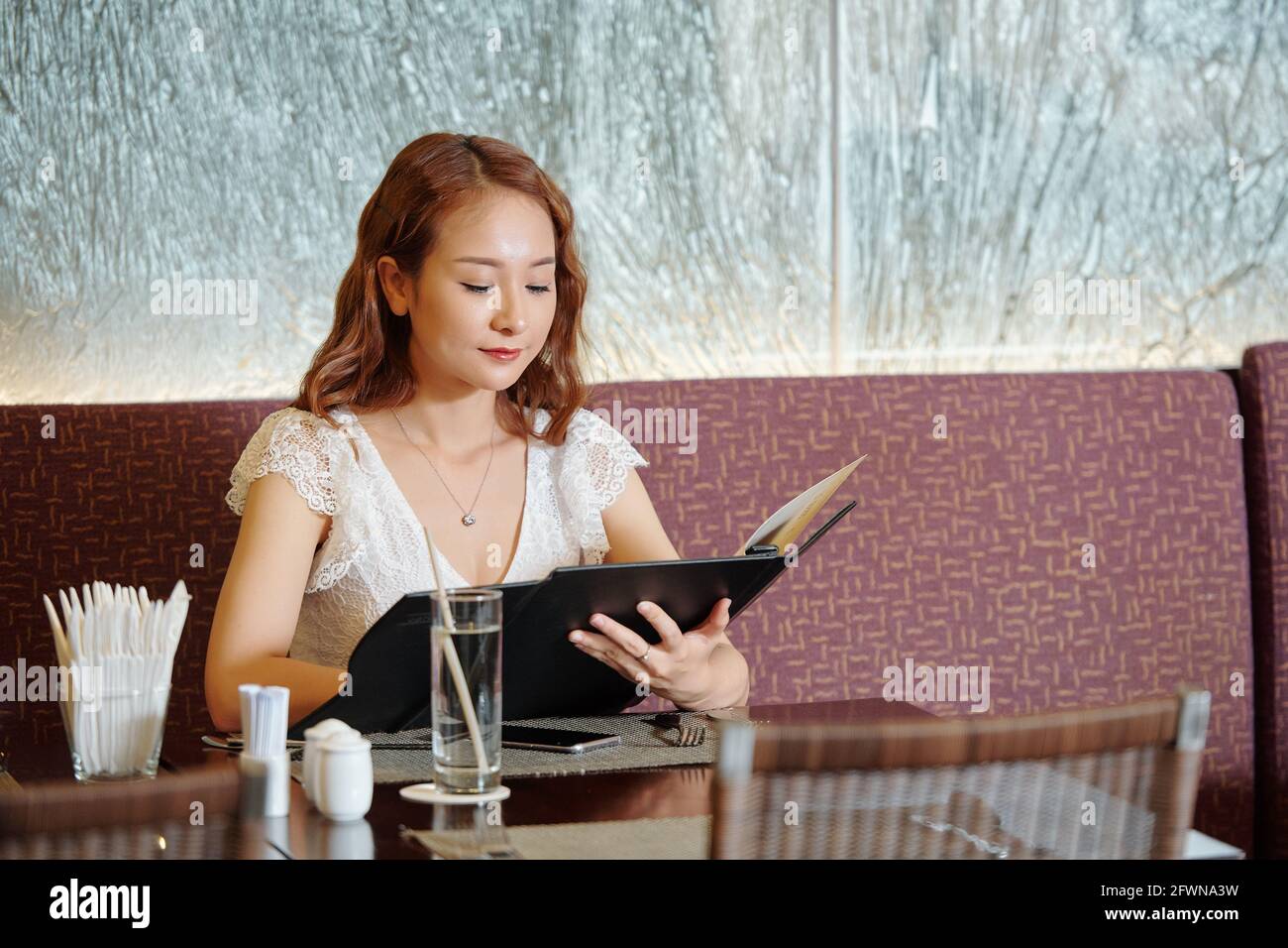 Pretty young woman sitting at restaurant table and reading menu deciding what to have for dinner Stock Photo