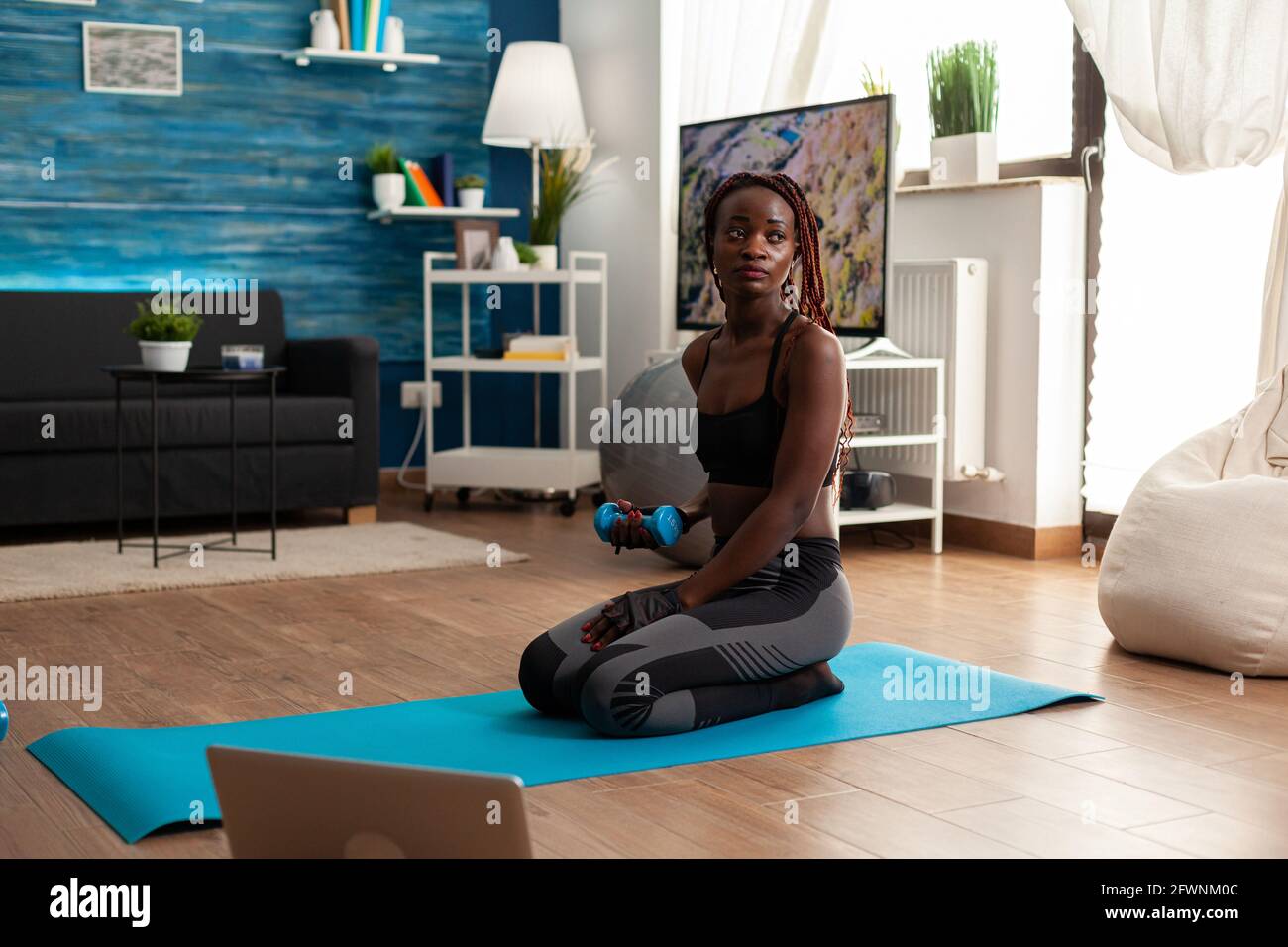 Fit athletic active black woman flexing arms working biceps, sitting on yoga mat in home living room exercising following video instructions from laptop, having a healthy lifestyle. Stock Photo