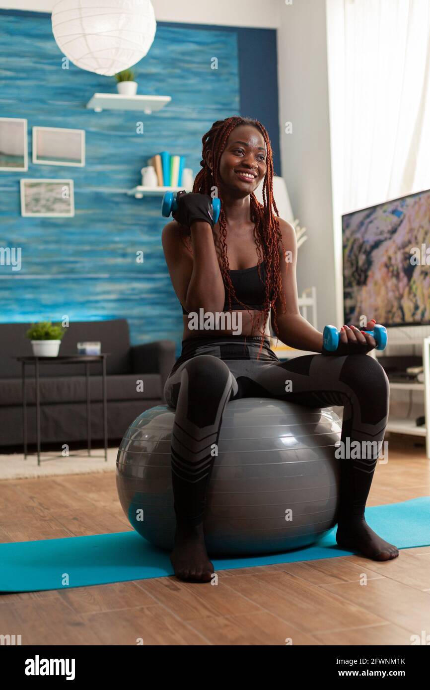 Smiling cheerful african woman flexing arm working out biceps, using dumbbells sitting on stability ball making training harder after warm up. Strong athletic person doing sports at home. Stock Photo