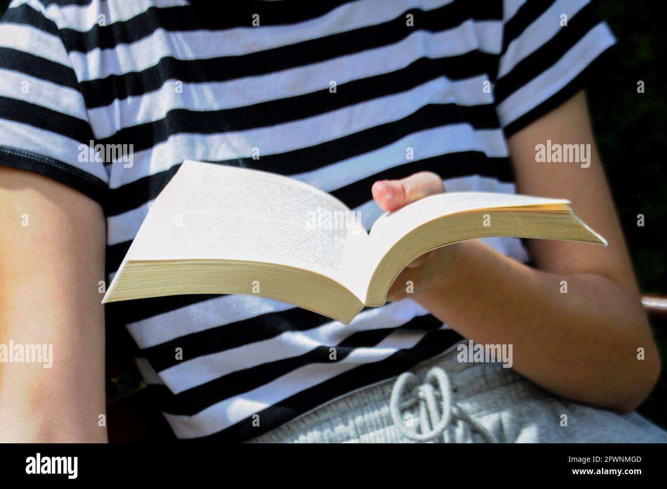 Person sitting on the bench, holding a white book and reading outdoors. Leisure time in the garden. Stock Photo