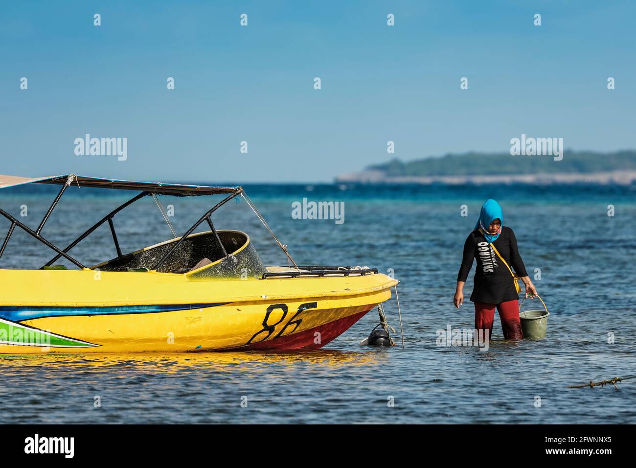 Local woman harvesting seaweed from the seashore by a tourist speedboat at this far South resort town beach; Tanjung Bira, South Sulawesi, Indonesia Stock Photo