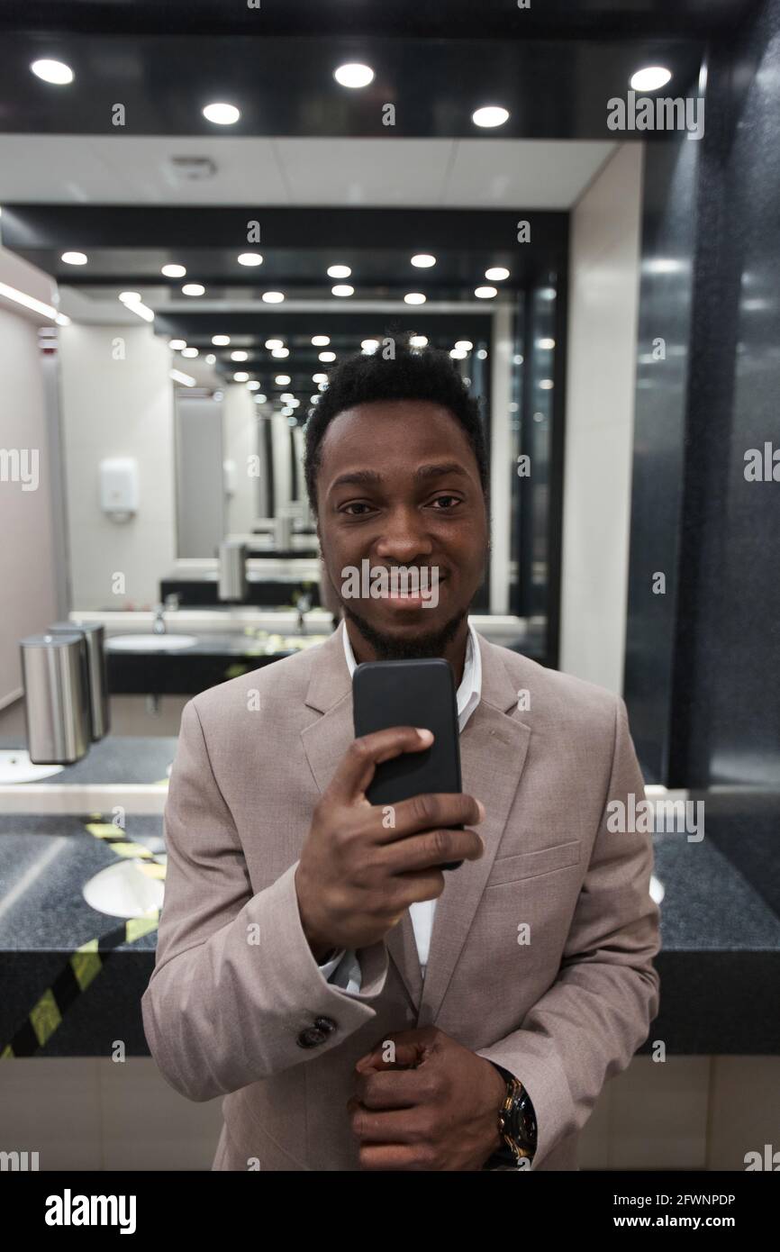 Vertical portrait of young African-American businessman taking mirror selfie in public restroom Stock Photo
