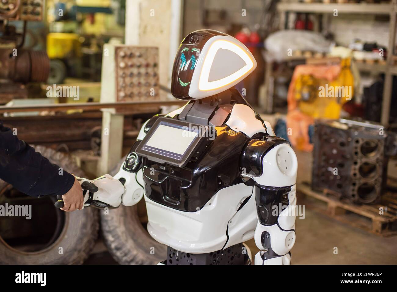 Humanoid robot on wheels with a monitor on his chest, shaking hands with a man. The old cluttered garage. Stock Photo