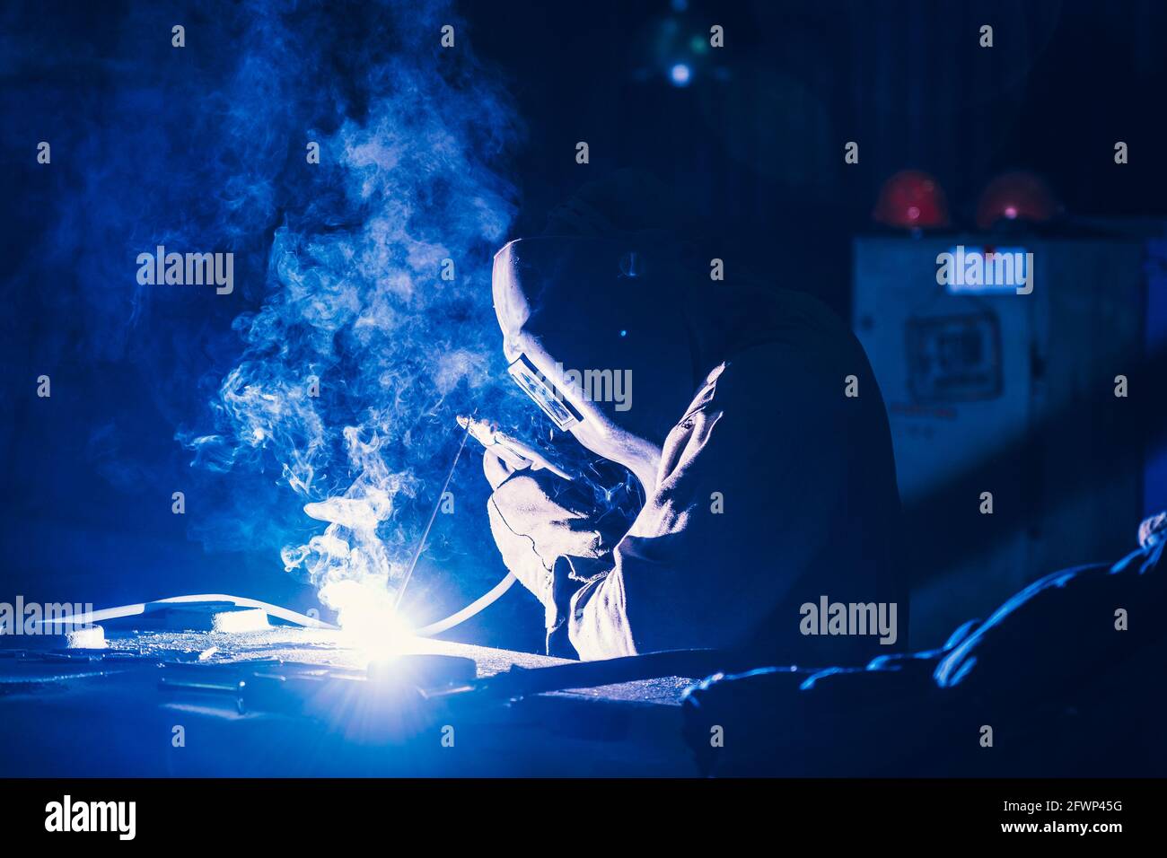 Welder in protective mask, sparks and smoke from welding flares, industrial background. Stock Photo