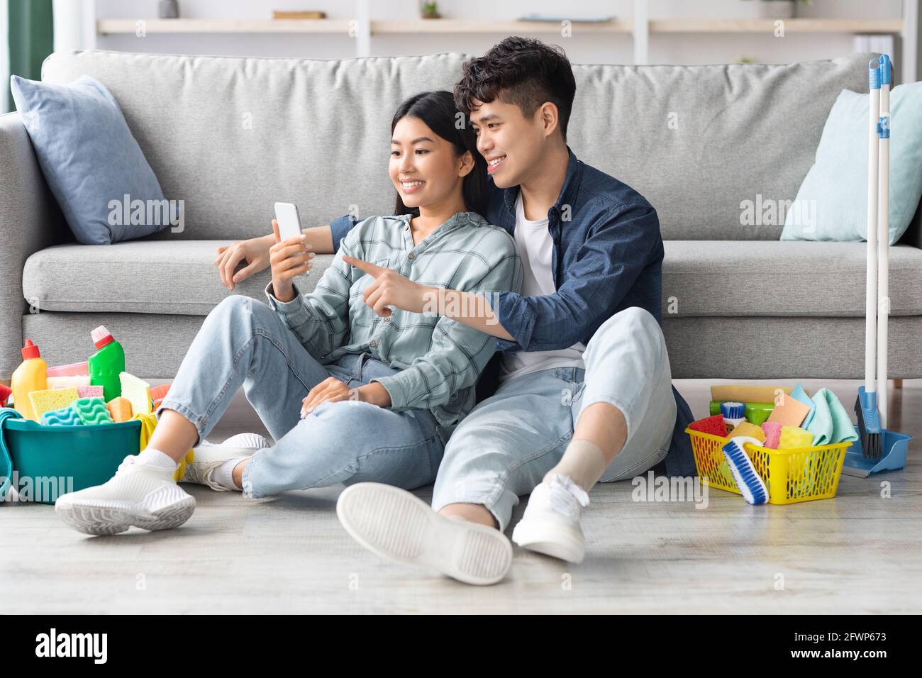 Loving chinese couple with cleaning tools resting, using smartphone Stock Photo
