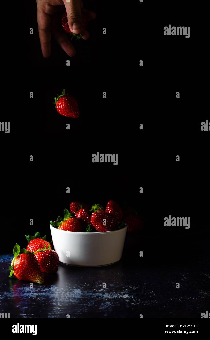 Frozen movement of a strawberry falling from the cook's hand into a bowl with more strawberries. Dark Food Photography Stock Photo