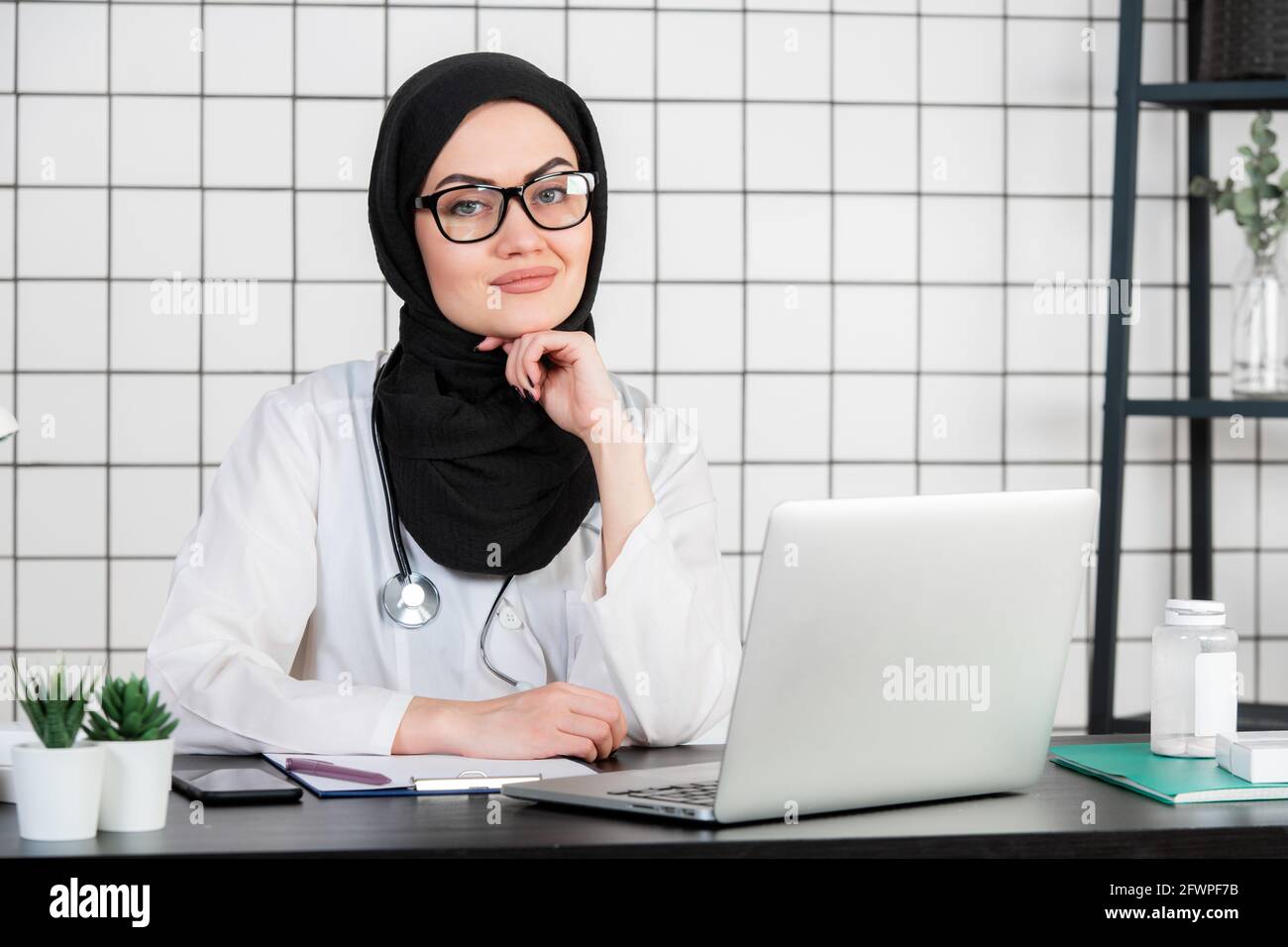 A Muslim Doctor smiling with glasses and stethoscope and documents on a white office background. Stock Photo