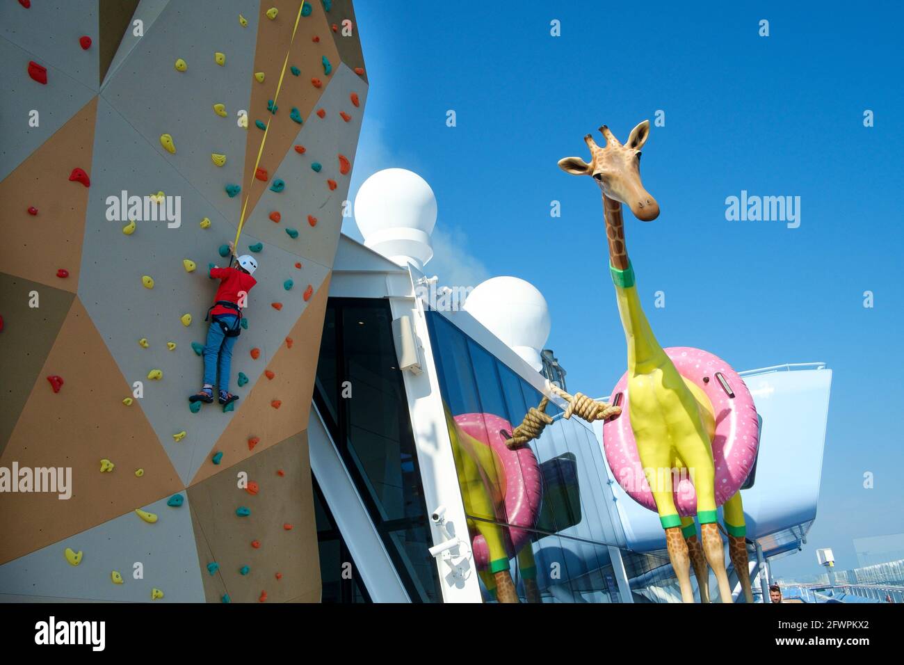 A young boy on the climbing wall next to a giant Giraffe called Gigi by french artist Jean Francois aboard Royal Caribbean ship Anthem of the Seas. Stock Photo
