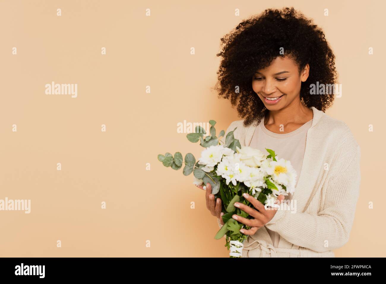 african american woman holding bouquet of daisies and smiling on beige ...