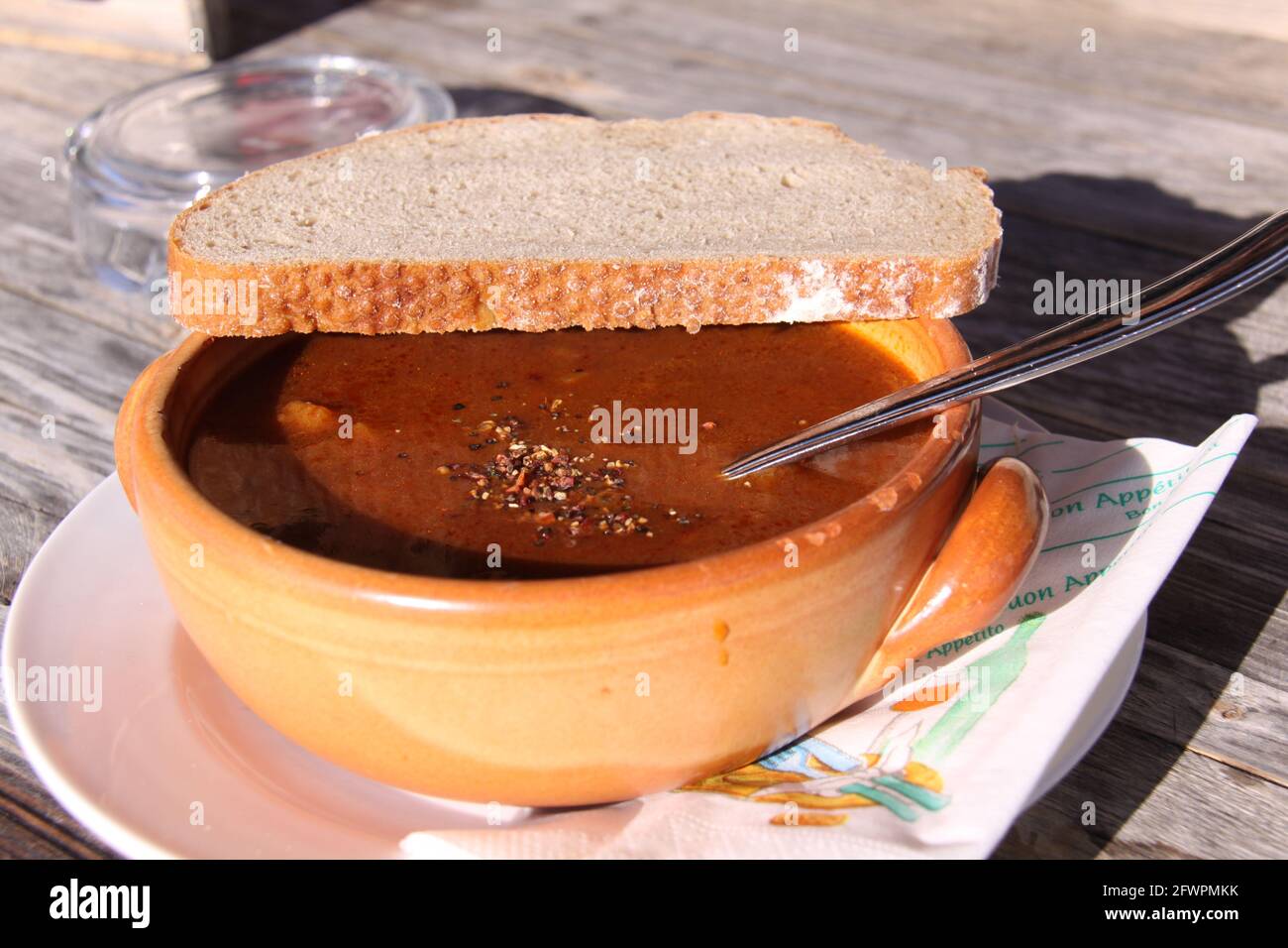 Rustic bowl of goulash soup with a slice of bread on top. (text on the napkin: 'enjoy your meal' in French and Italian) Stock Photo