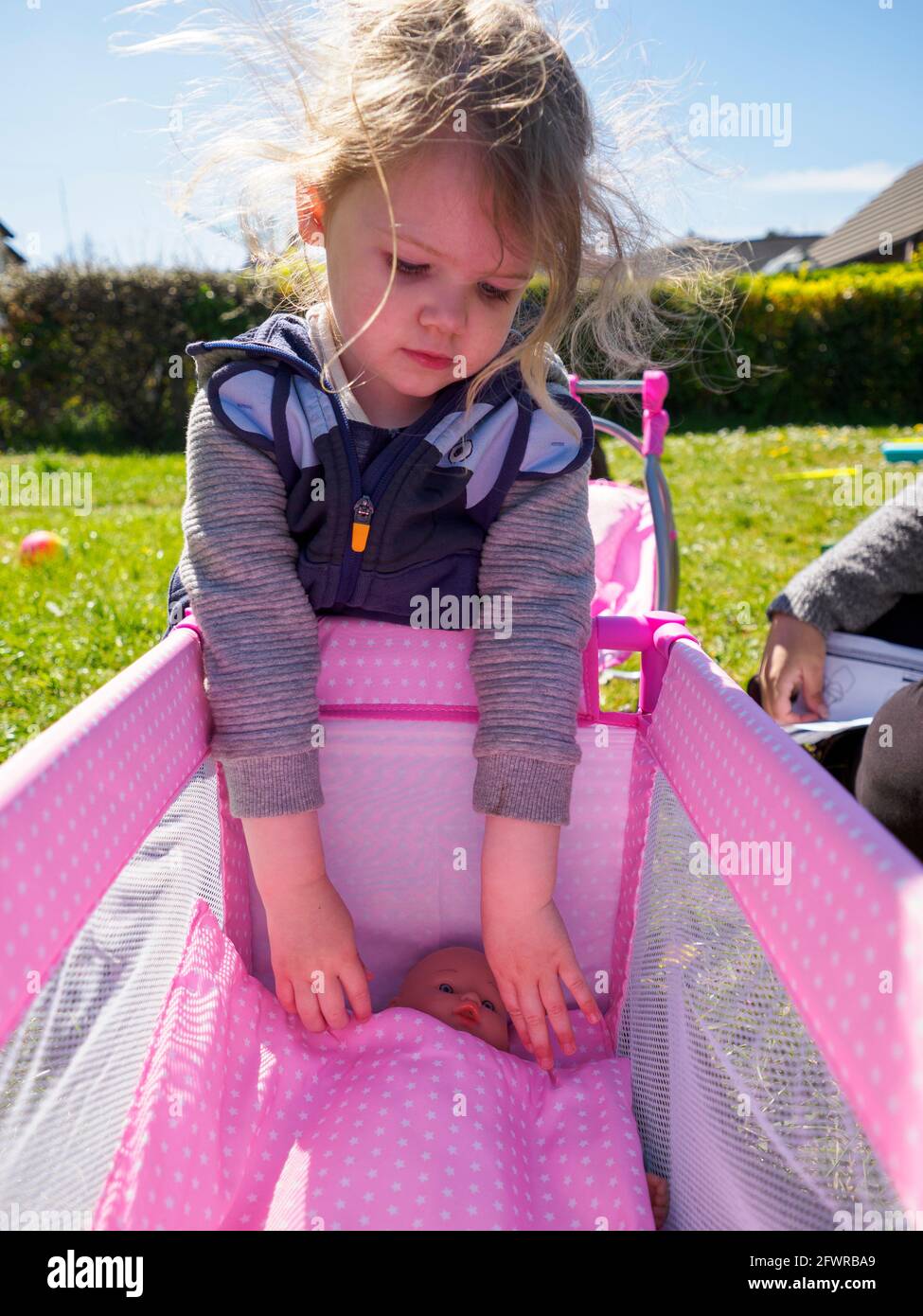 Toddler playing with a baby doll in a toy cot in the garden, UK Stock Photo