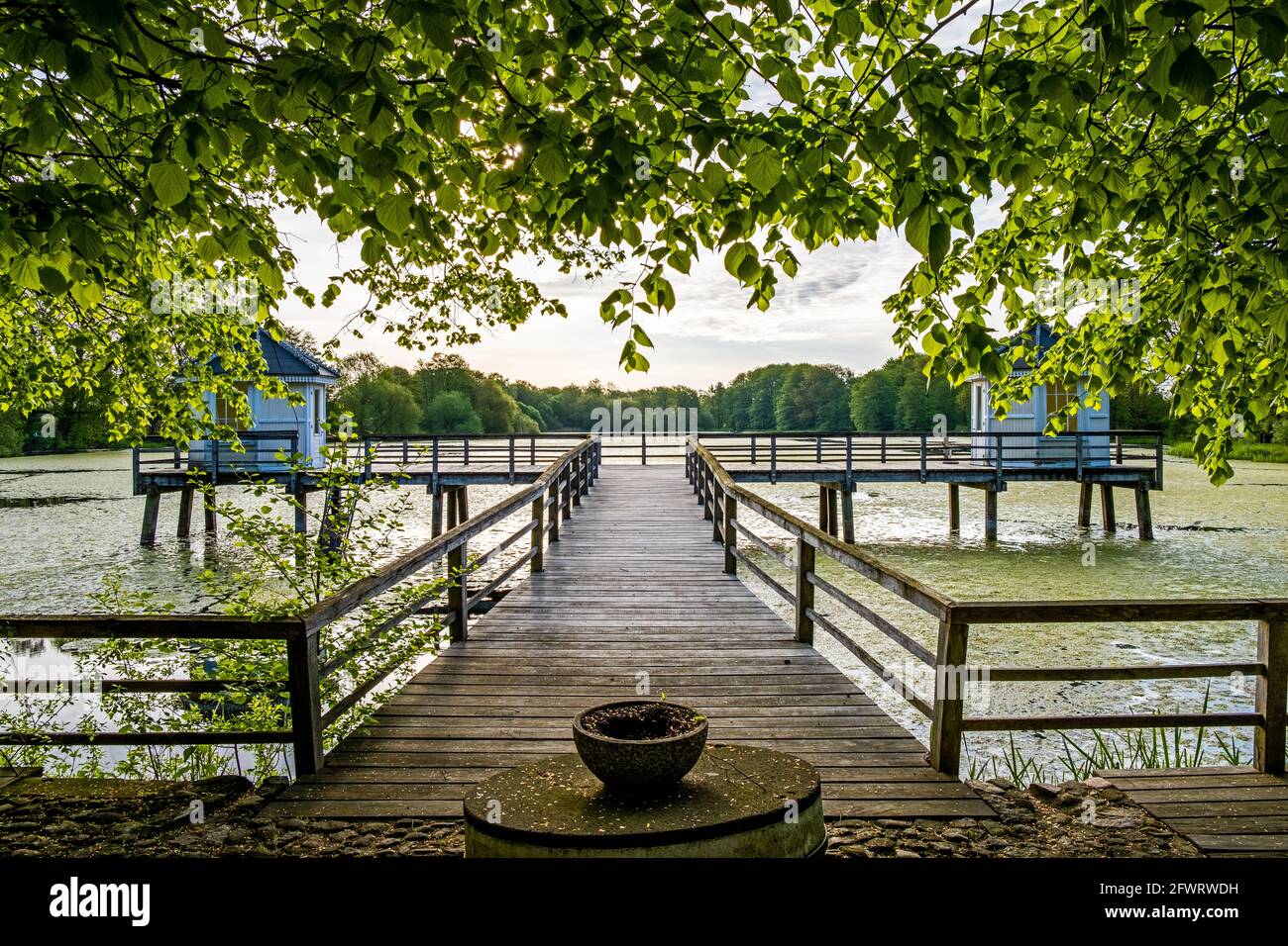 Landscape photography of a historic wooden bridge leading to two symmetric bath houses at a lakeside with greenish water surface and leafage Stock Photo