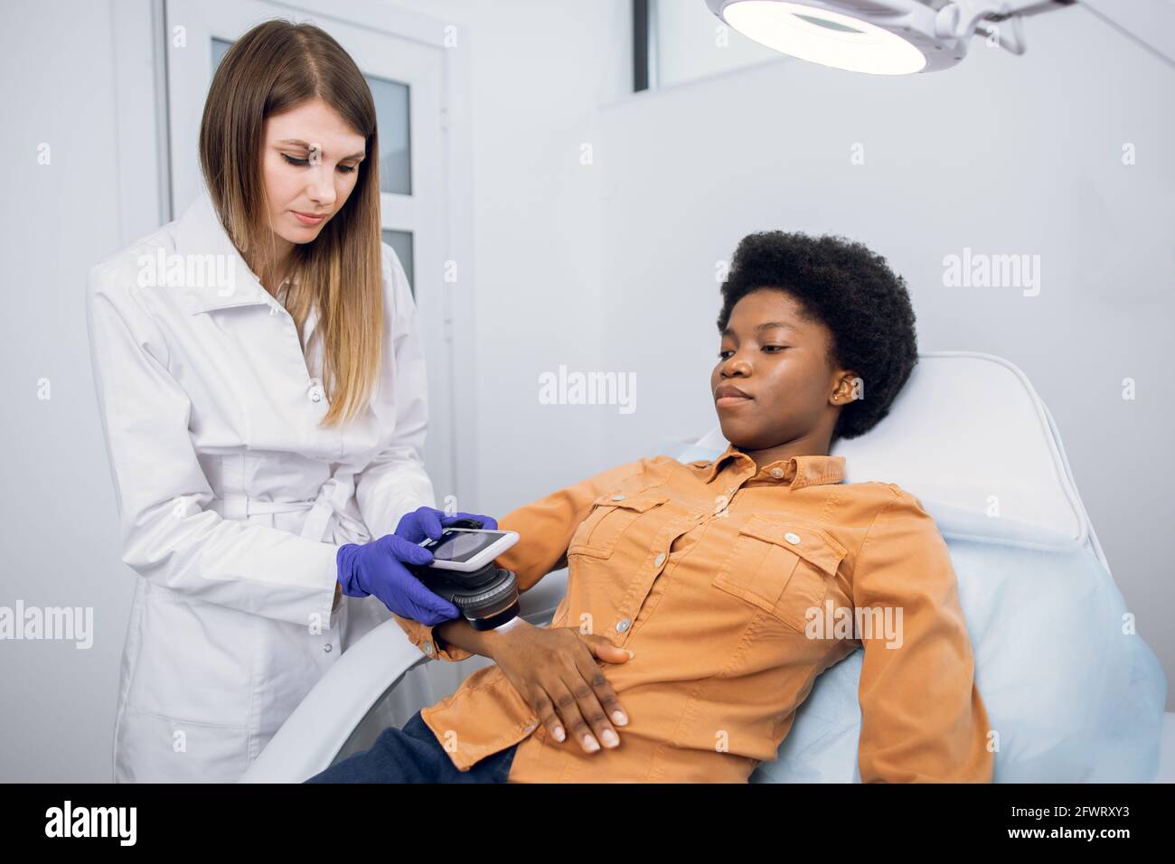 Skin cancer and melanoma prevention. Professional doctor dermatologist examines the patient's moles on arm of African lady with the help of a modern device for the dematoscopy Stock Photo