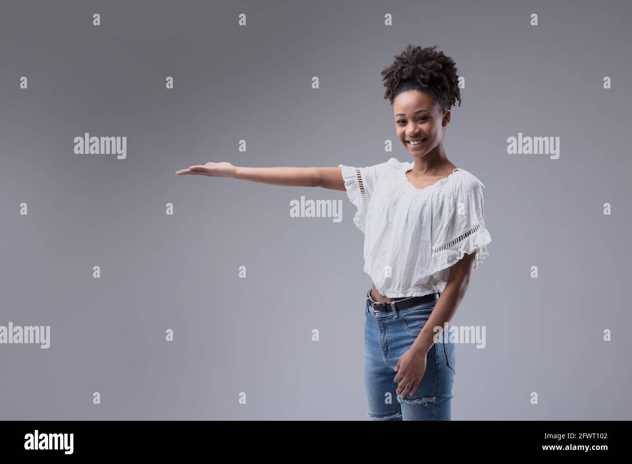 Attractive young Black girl in jeans holding out her empty palm for product placement for advertising with a happy friendly confident smile on a grey Stock Photo