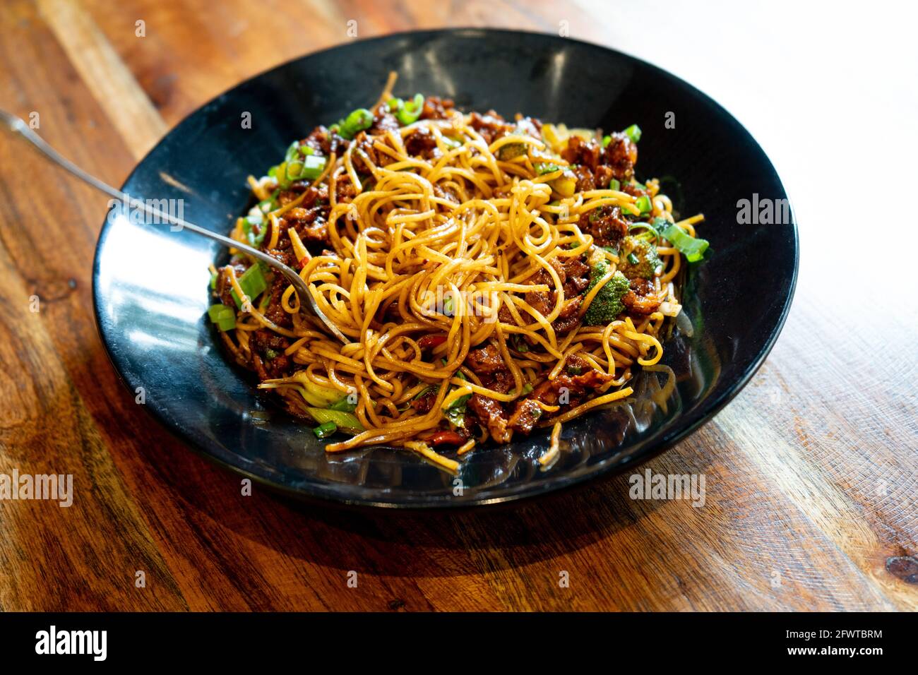 Indo chinese thin noodles garnished with green vegetables and spices with chiceken tofu and paneer placed on a black bowl on a wooden table Stock Photo