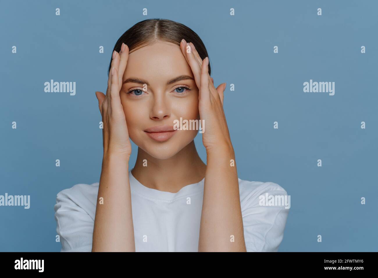 Young brunette female model touches face enjoys softness of skin has natural makeup dressed in casual white t shirt looks calmly at camera isolated Stock Photo