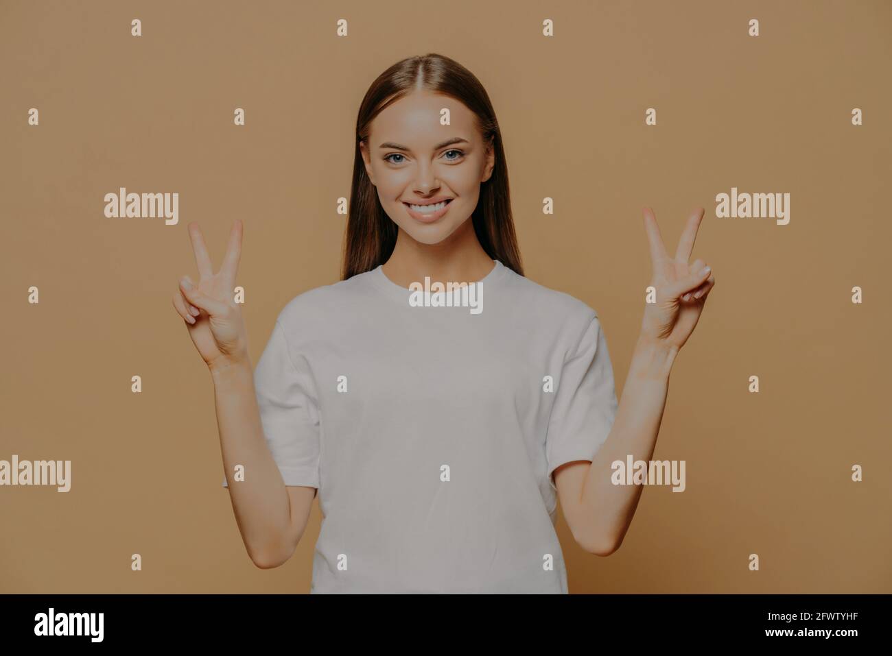 Happy optimistic young Caucasian woman raises arms shows peace gesture with fingers does victory sign says number two wears casual white t shirt Stock Photo