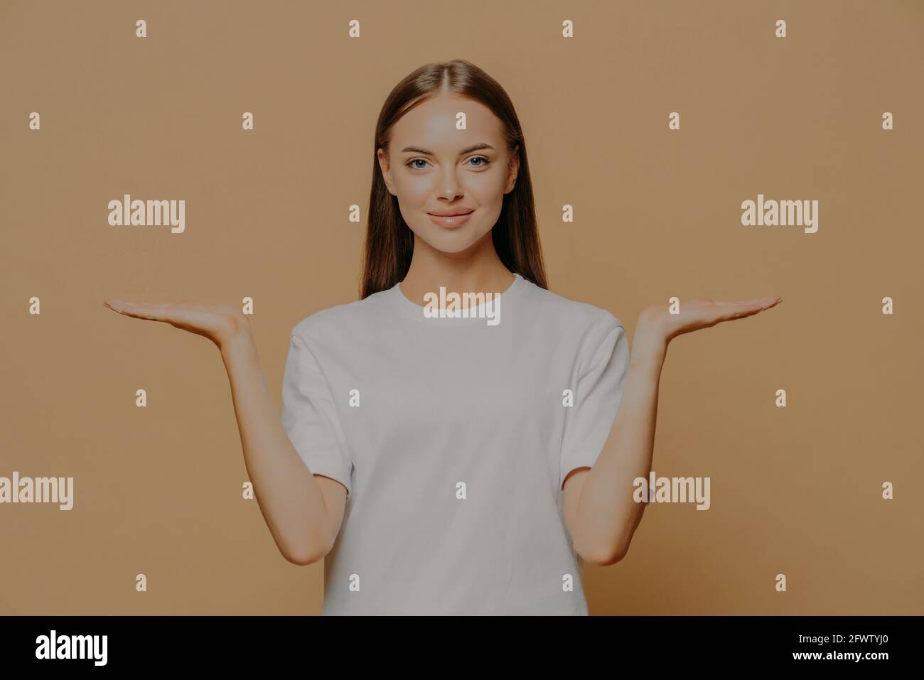 Woman raises palms pretends holding something over blank copy space against brown background wears casual white t shirt proposes product Stock Photo
