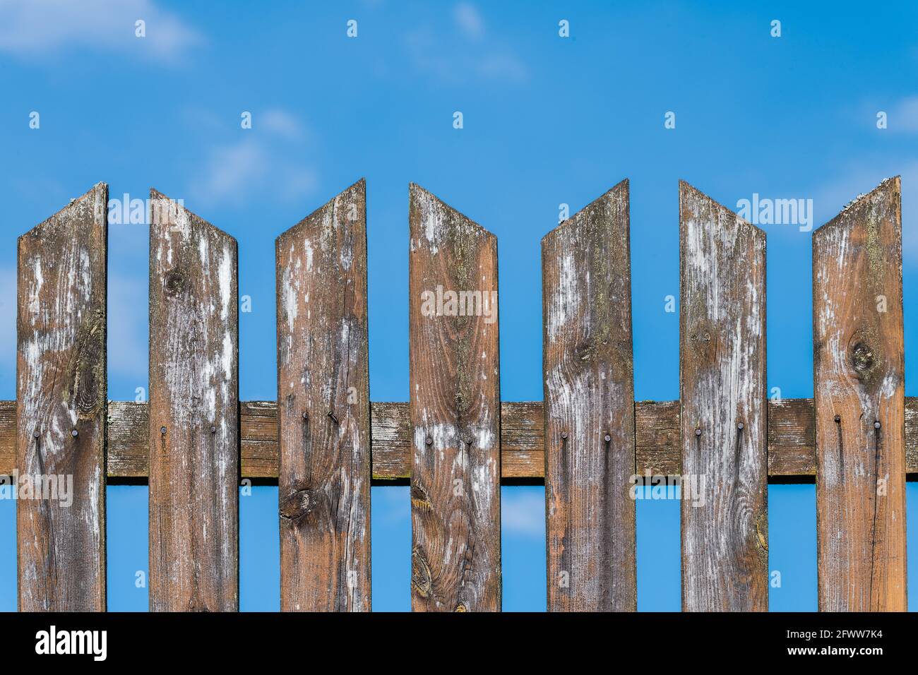 Top of old weathered pointed wood fence on a blue sky background. Shabby wooden planks, nature grains or knots, Pale smudges of white paint remainders. Stock Photo