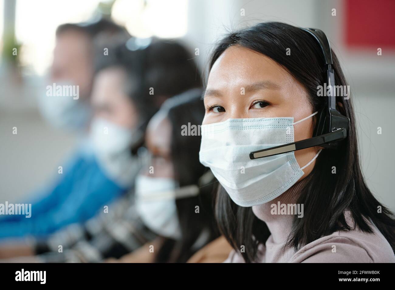 Portrait of Asian customer service operator in mask and wireless headset with microphone sitting against colleagues in call center Stock Photo