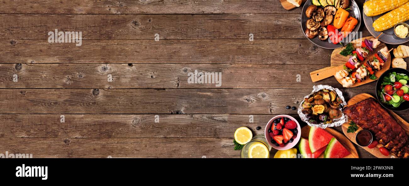Summer BBQ or picnic food corner border over a rustic wood banner background. Assorted grilled meats, vegetables, fruits, salad and potatoes. Overhead Stock Photo