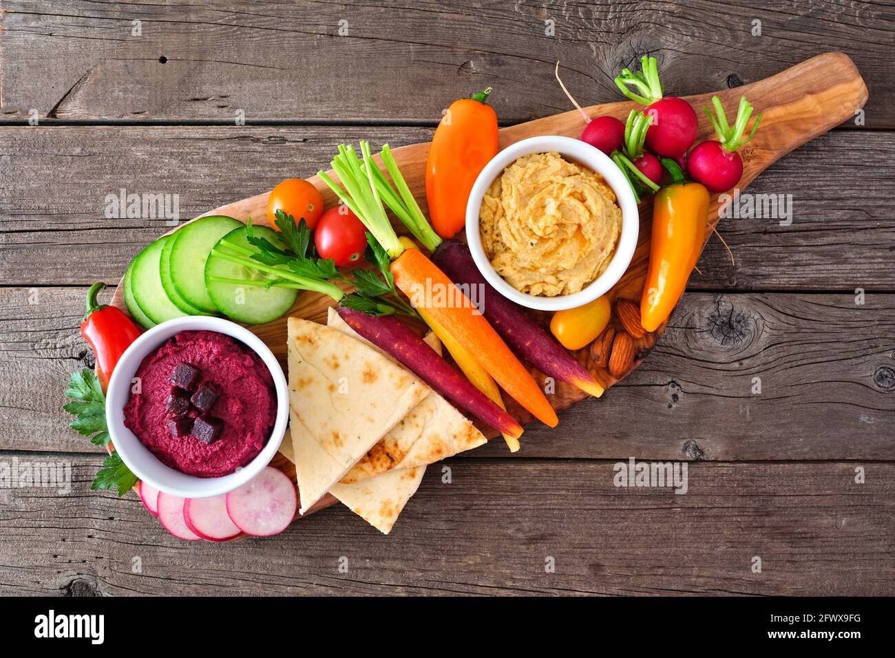 Assortment of fresh vegetables and hummus dip on a serving board. Top view on a rustic wood background. Stock Photo