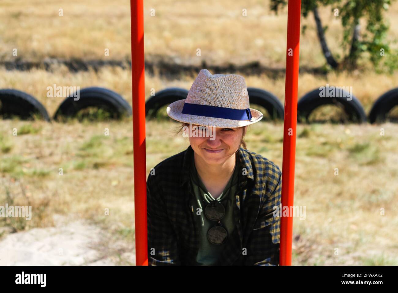 Defocus smiling young woman in checkered shirt and hat swinging on swing on playground. Hipsters and hippie portrait. Countryside area. Childlike adul Stock Photo