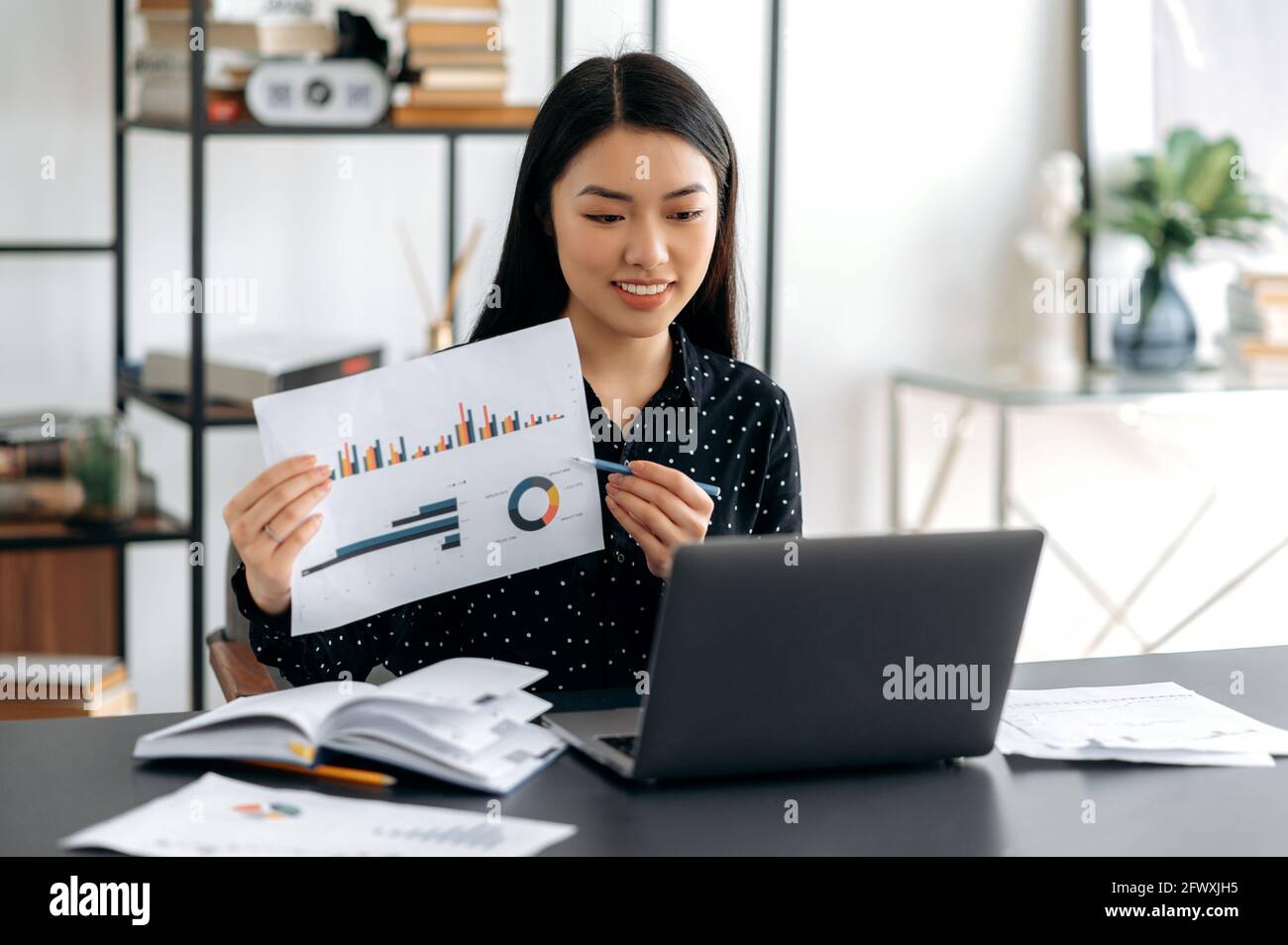 Young adult friendly smiling confident asian woman in formal shirt, sitting at her work desk, analyzing report, showing financial graph to colleagues during communicating by video conference using Stock Photo