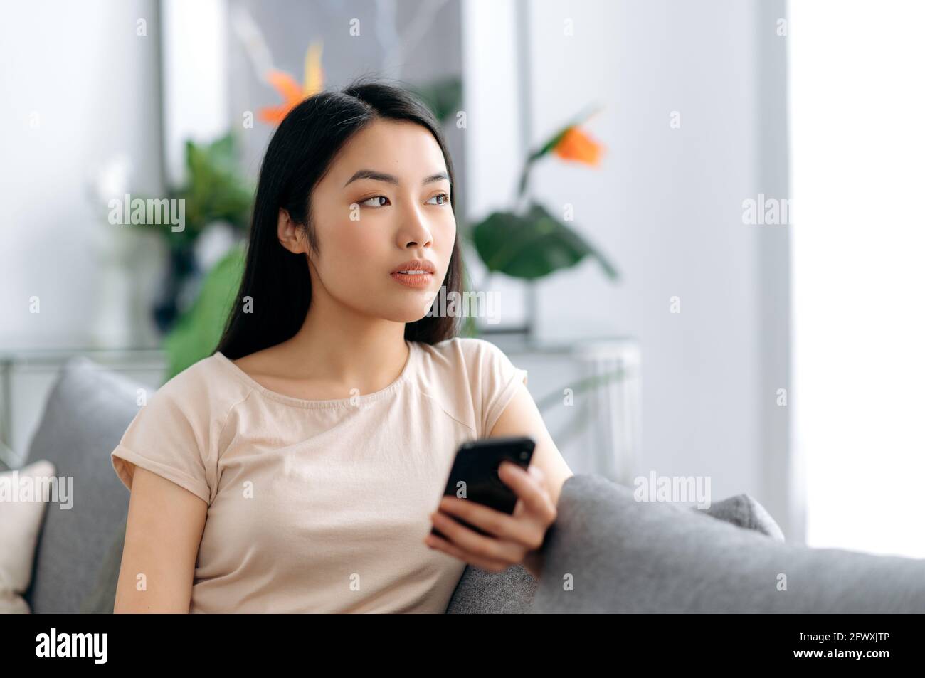 https://c8.alamy.com/comp/2FWXJTP/pensive-young-asian-beautiful-woman-uses-her-mobile-phone-while-sitting-on-the-sofa-in-casual-clothes-browses-the-internet-and-social-networks-texting-with-friends-or-family-looks-away-thoughtfully-2FWXJTP.jpg