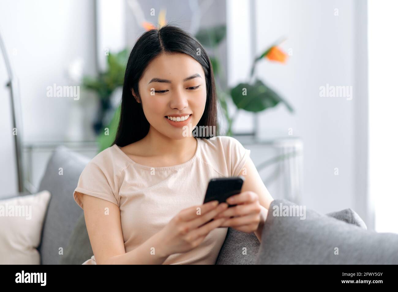 Joyful japanese attractive girl using her smartphone while sitting on sofa in stylish clothes, browsing internet and social networks, texting with friends or family, found out good news, smiles Stock Photo