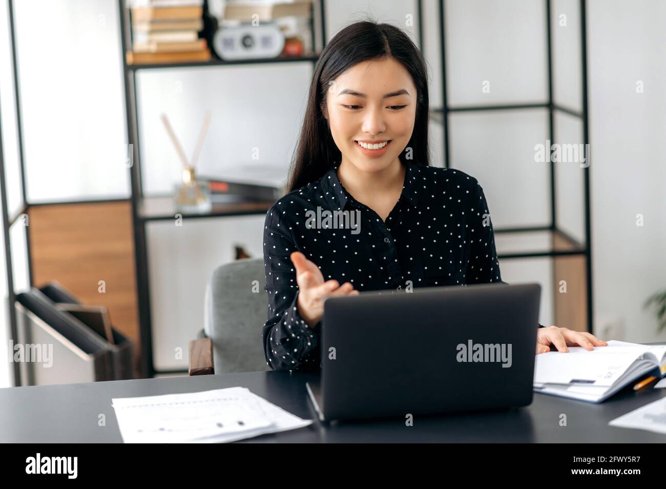 Charismatic successful confident Asian business lady, freelancer, manager, using laptop, talking on video conference with client or colleague, discussing business project, gesturing with hand, smiling Stock Photo