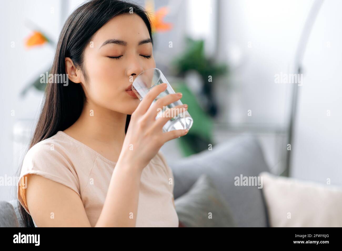 Pretty young asian woman drinks a glass of water while sitting on the sofa ...