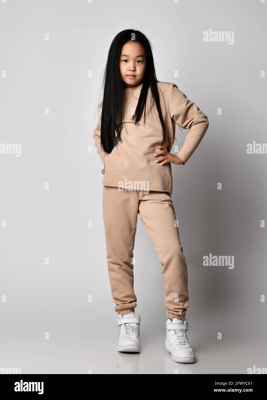cool asian kid girl green pants hoodie sweater and sneakers stands holding hand at waist posing in new sportswear 2FWYC61