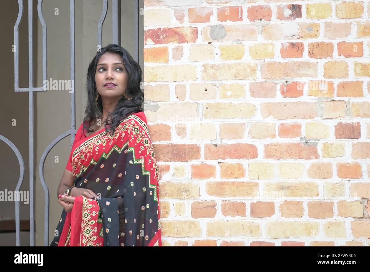 Portrait of a beautiful and smiling Indian woman wearing red saree. Standing next to brick wall, outdoor setting. Stock Photo