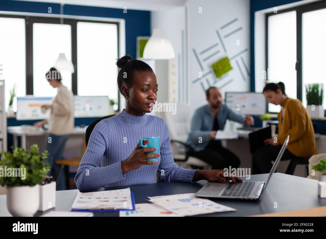 African businesswoman holding cup of coffie reading project briefing in start up workplace. Diverse team of business people analyzing company financial reports from computer. Stock Photo