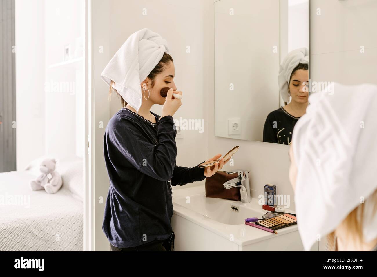 Side view of young Hispanic lady in casual clothes and towel on head applying foundation on face with brush while standing in front of mirror near unr Stock Photo