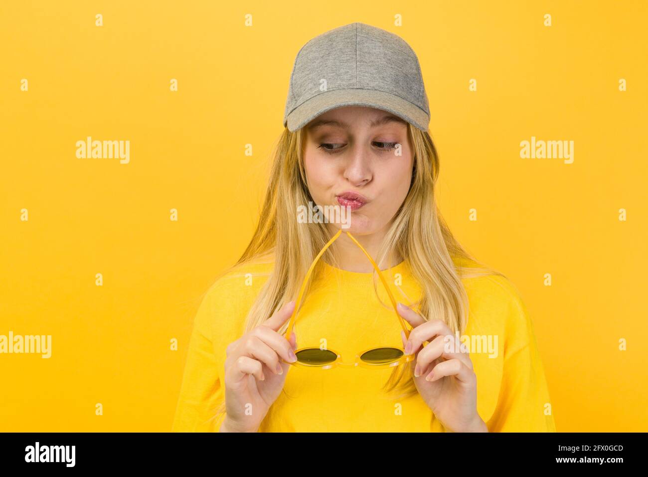 Perplexed Woman in yellow sweater and cap holding stylish sunglasses while standing on yellow background Stock Photo